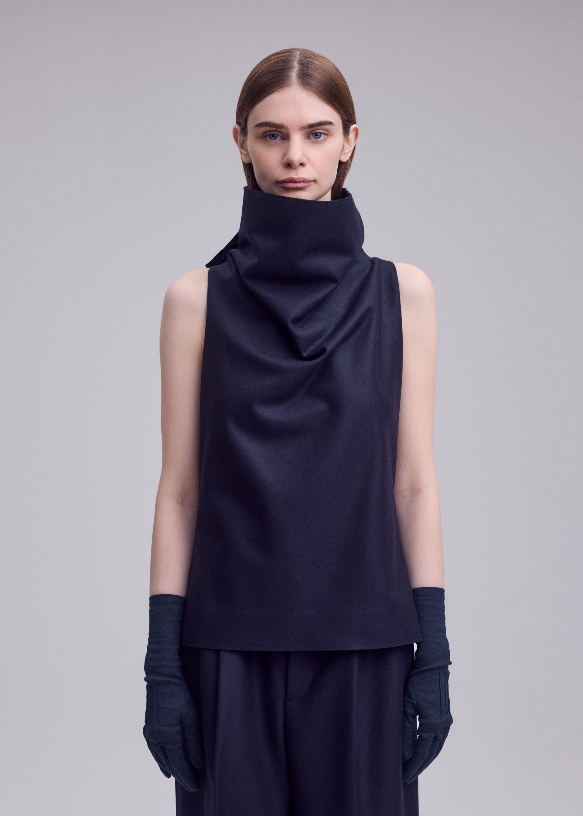 Zoomed view A person with straight brown hair wears the ISSEY MIYAKE SHELL sleeveless jersey top, paired with black pants and black gloves, standing against a plain light gray background in signature ISSEY MIYAKE style.