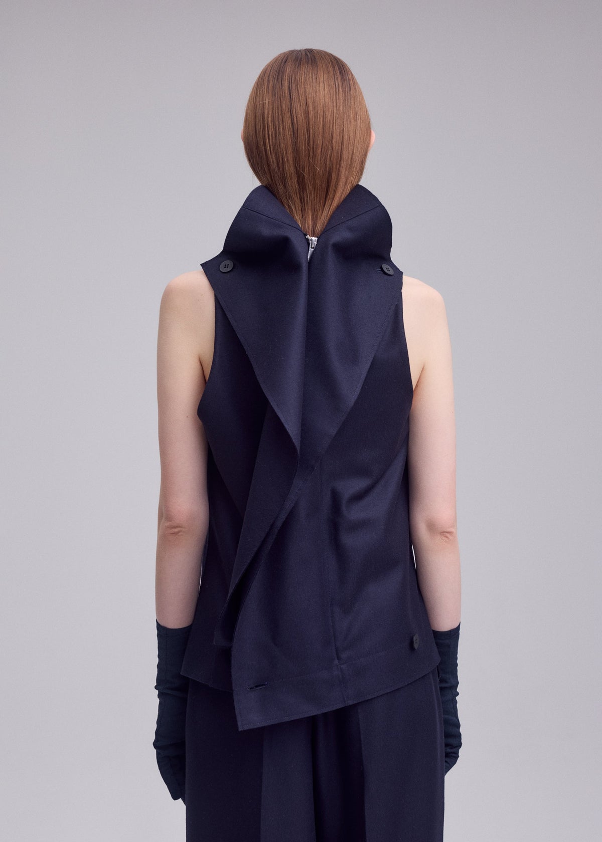 A person with straight, long hair stands facing away, wearing ISSEY MIYAKE’s SHELL—a dark sleeveless top crafted from soft jersey material featuring layered, draped fabric—and dark gloves. The plain light gray background evokes the understated elegance characteristic of ISSEY MIYAKE designs. view 8 of 8