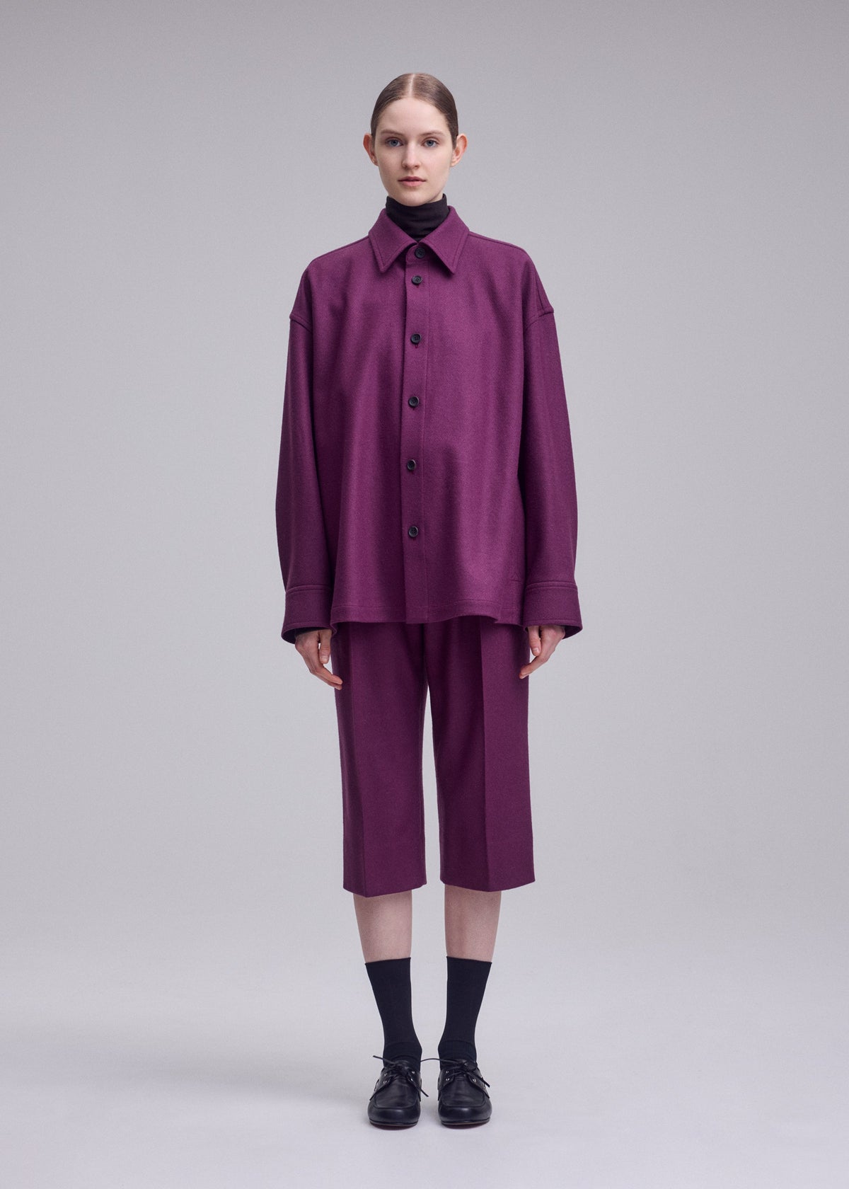 A person stands against a plain background wearing the ISSEY MIYAKE MANNER loose-fitting purple regular collar shirt and matching knee-length shorts, paired with black socks and shoes. view 3 of 5
