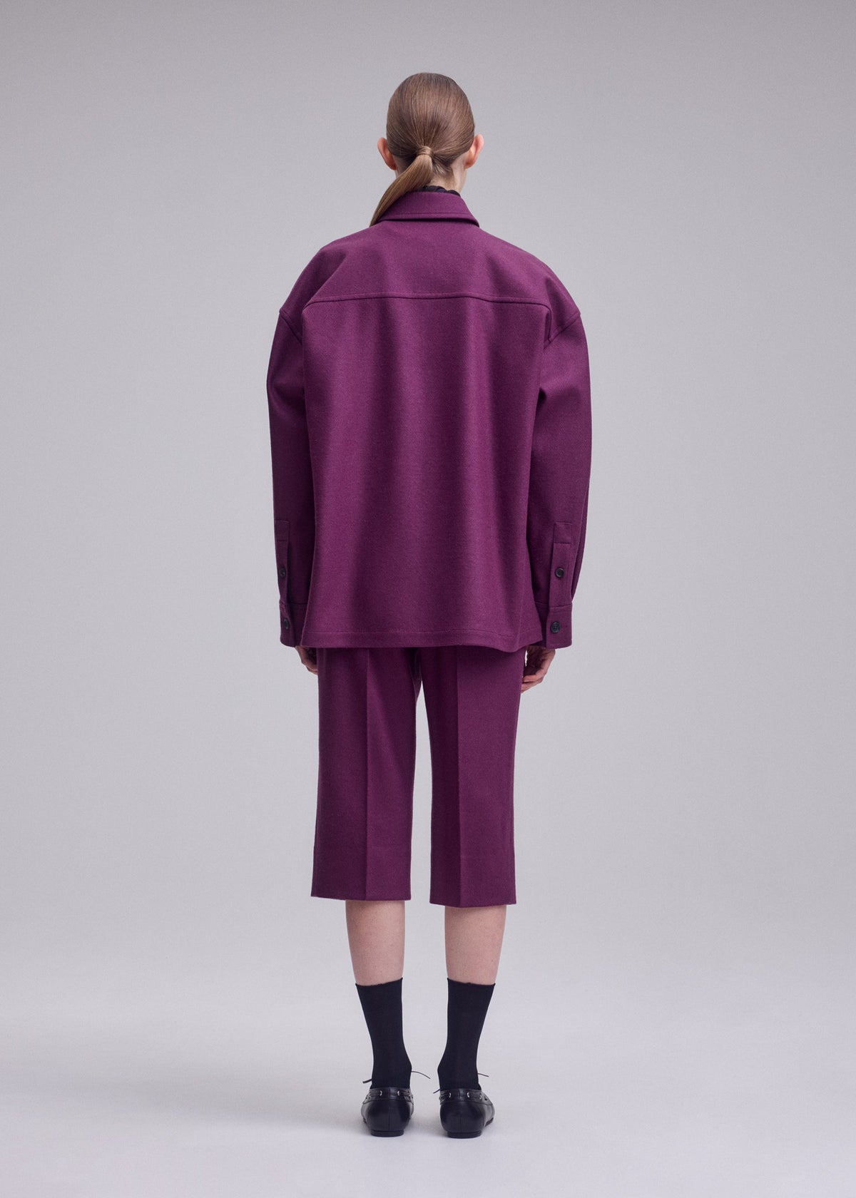 A person faces away, wearing ISSEY MIYAKE’s MANNER—a loose purple jacket and matching knee-length shorts—paired with black socks and shoes. Their hair is neatly pulled back against a plain, neutral background. view 4 of 5