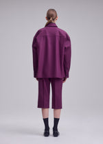 A person faces away, wearing ISSEY MIYAKE’s MANNER—a loose purple jacket and matching knee-length shorts—paired with black socks and shoes. Their hair is neatly pulled back against a plain, neutral background.