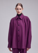 A woman with straight brown hair in a low ponytail wears ISSEY MIYAKE’s MANNER—an oversized purple shirt made from leftover fabric—over matching pants and a black turtleneck, against a plain light gray background.