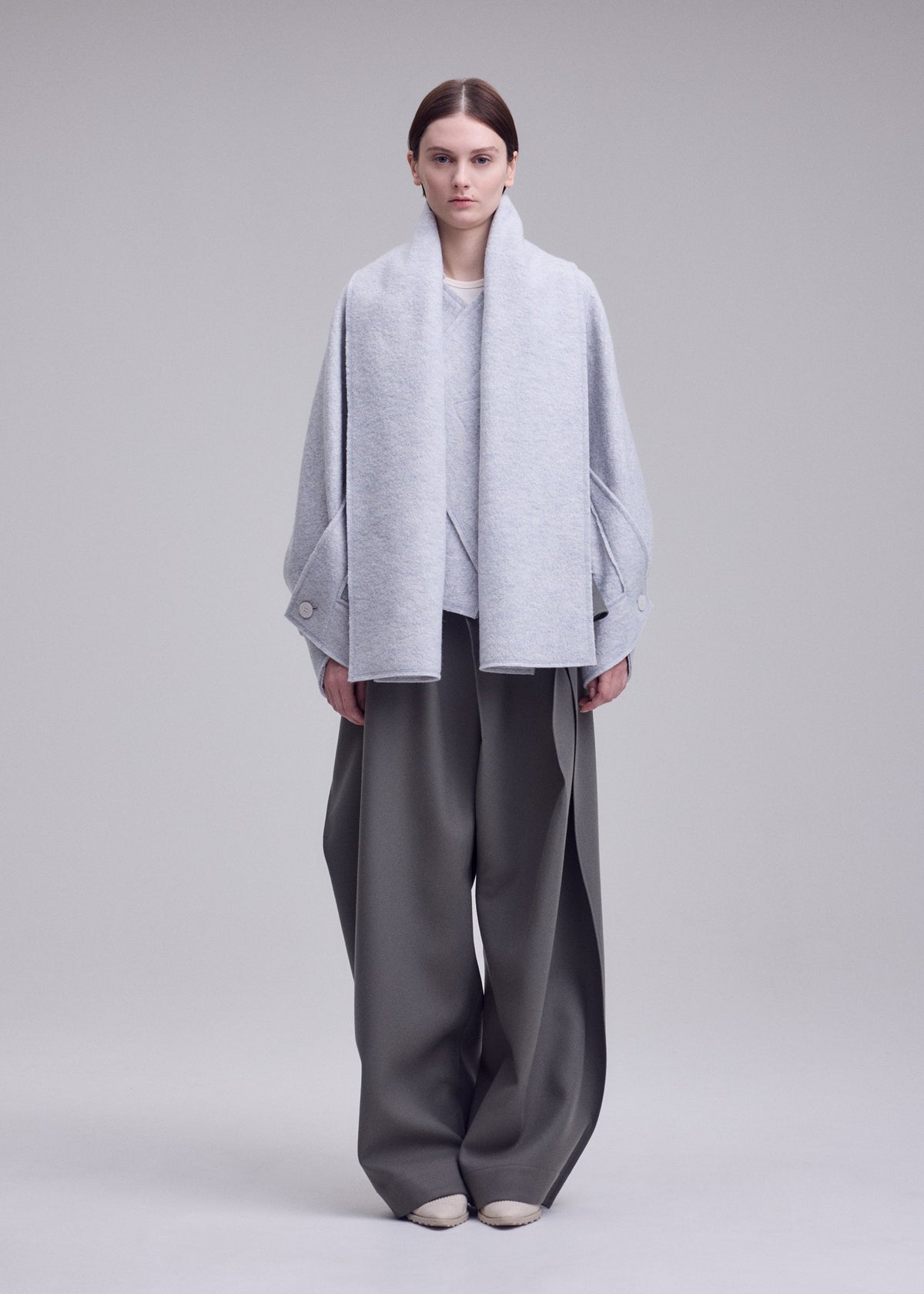 A person stands against a gray background wearing ISSEY MIYAKE’s WOOLEN GRANITE merino wool coat with detachable scarf, wide-legged recycled polyester gray pants, and light-colored shoes. view 4 of 7