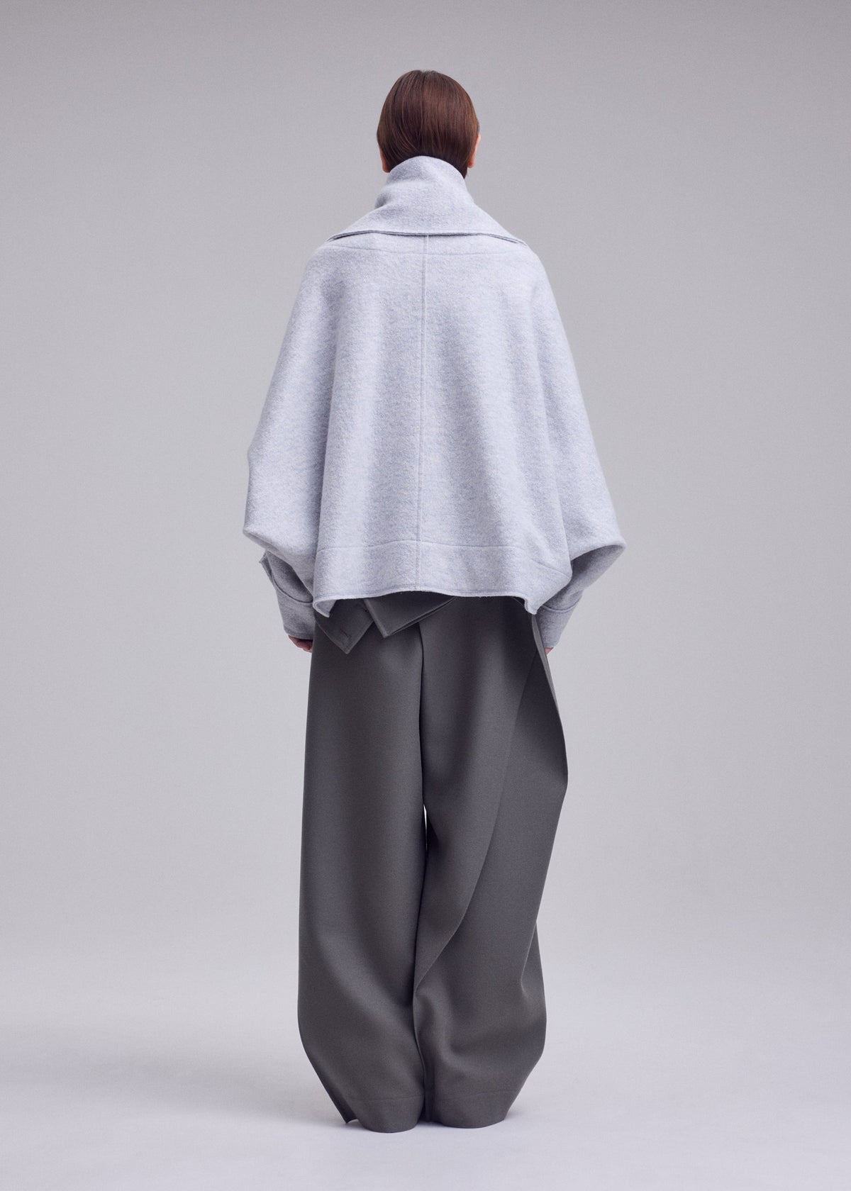 Person stands with back to camera wearing ISSEY MIYAKE’s WOOLEN GRANITE, a light gray oversized merino wool coat with detachable scarf, paired with wide-leg dark gray pants against a plain background. view 5 of 7