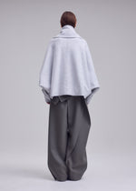 Person stands with back to camera wearing ISSEY MIYAKE’s WOOLEN GRANITE, a light gray oversized merino wool coat with detachable scarf, paired with wide-leg dark gray pants against a plain background.