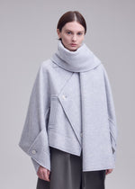 A person with short brown hair models the WOOLEN GRANITE by ISSEY MIYAKE—a light gray, asymmetrical merino wool coat with a large collar and buttons—over dark pants against a plain light gray background.