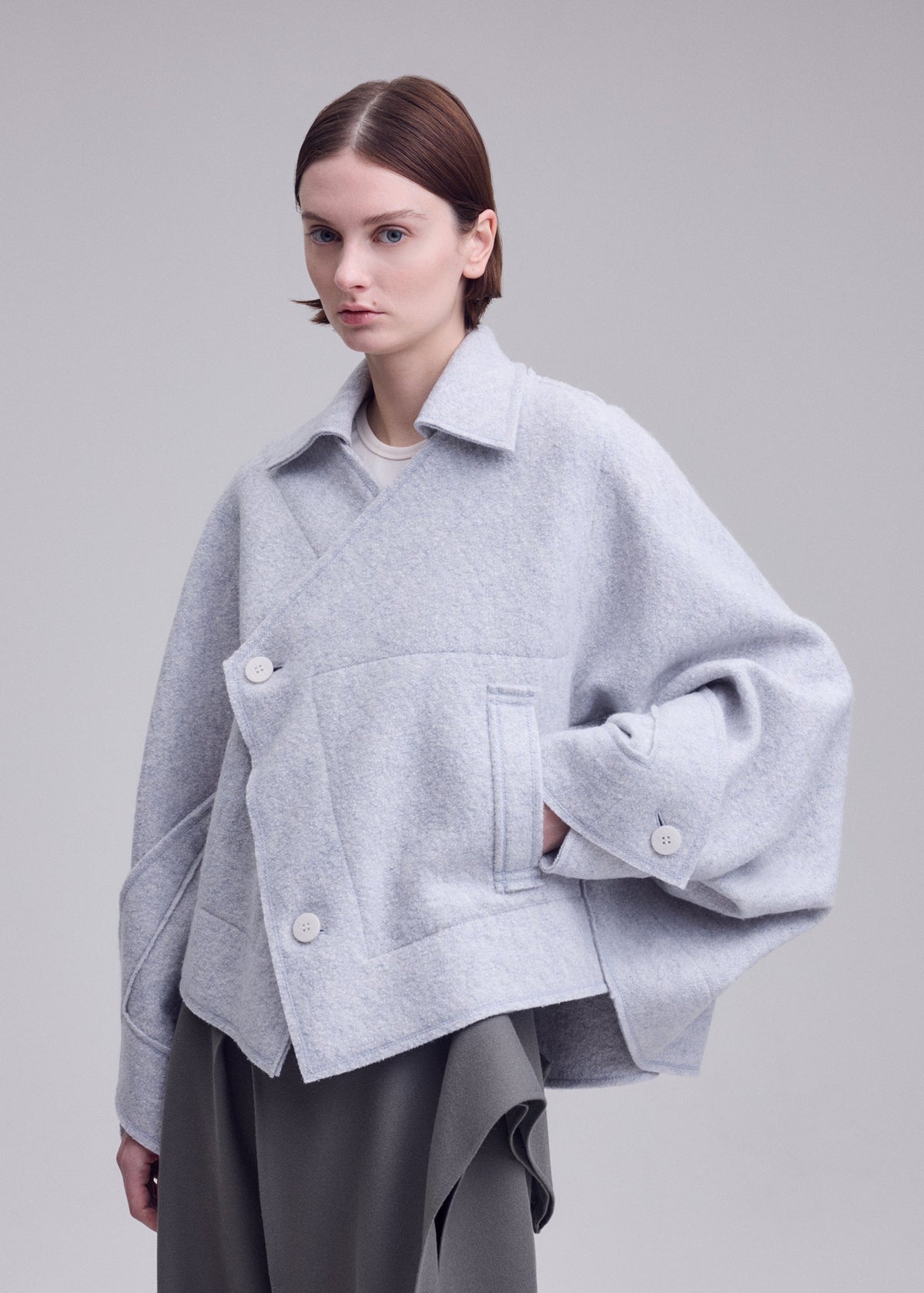 Zoomed view A woman with short brown hair models the ISSEY MIYAKE WOOLEN GRANITE: a loose, light gray merino wool coat with large buttons and wide sleeves, styled over a gray skirt against a plain background.