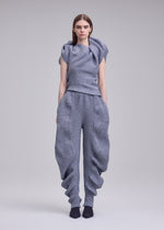 A person with straight brown hair faces forward against a plain light grey background, wearing ISSEY MIYAKE's FOUND STONE: a textured grey sculptural top and loose pants featuring a unique, organic silhouette.