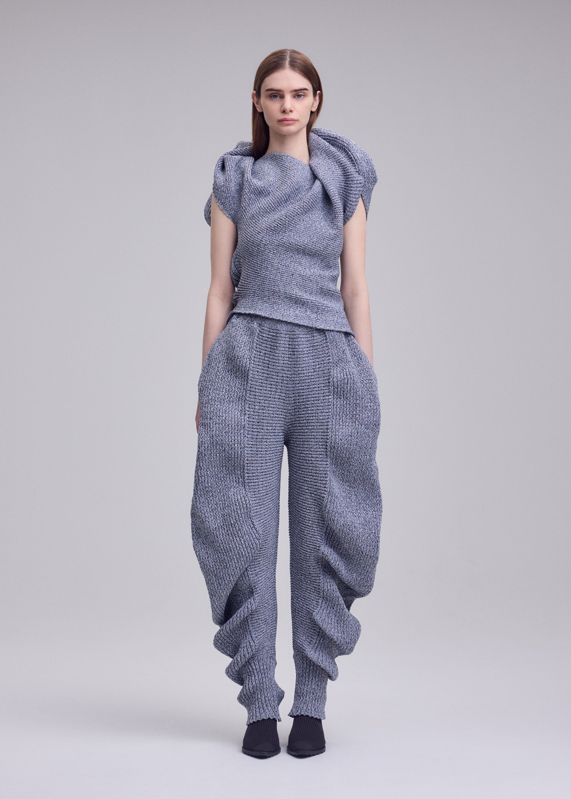 Zoomed view A person with straight brown hair faces forward against a plain light grey background, wearing ISSEY MIYAKE's FOUND STONE: a textured grey sculptural top and loose pants featuring a unique, organic silhouette.