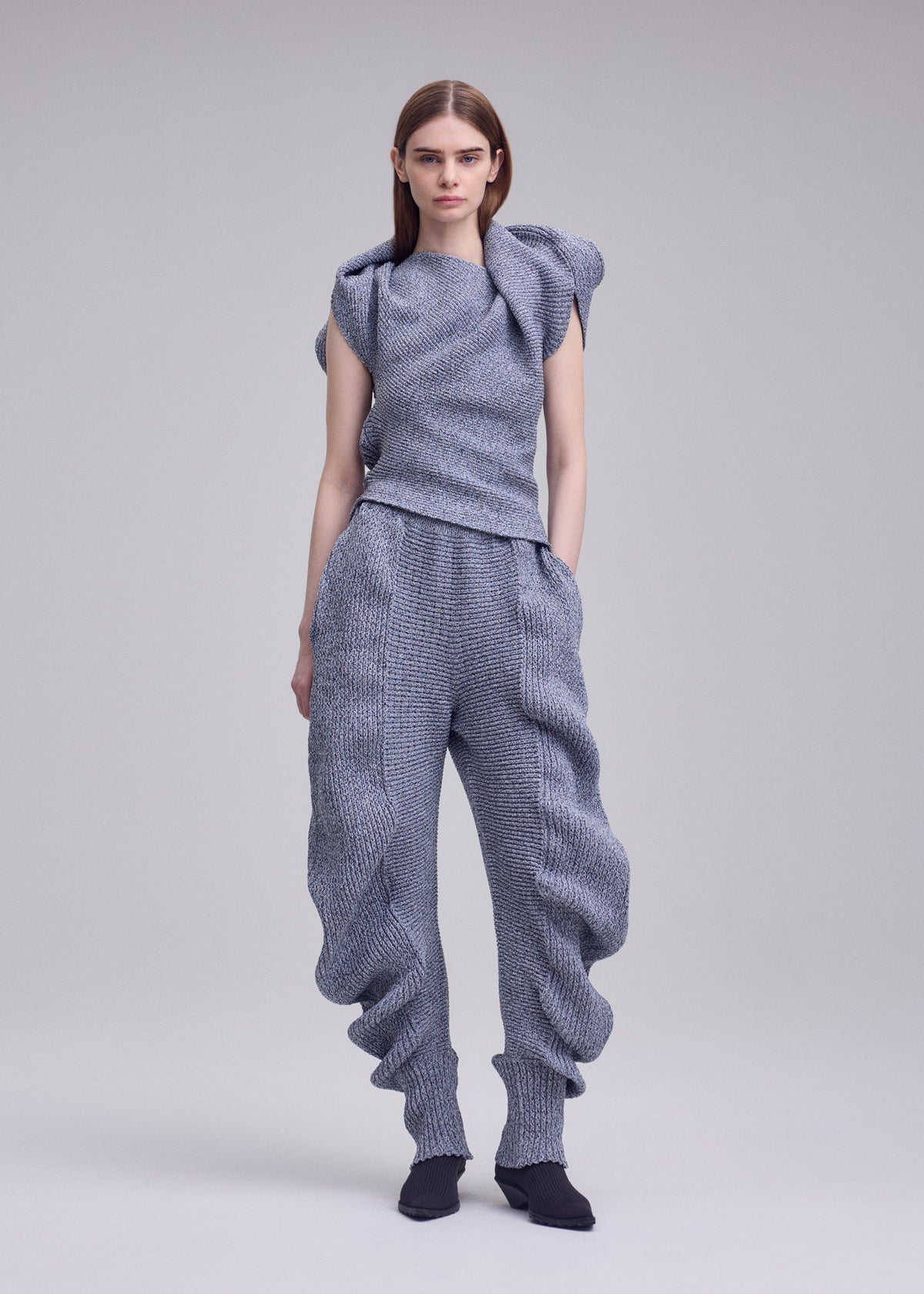 A woman with straight brown hair wears the ISSEY MIYAKE FOUND STONE outfit—textured, light gray, with an asymmetrical top and organic form pants—paired with black pointed shoes, standing against a plain background. view 7 of 8