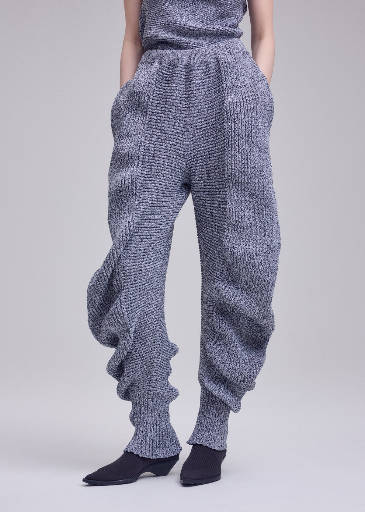 A person wears ISSEY MIYAKE's FOUND STONE chunky, textured knit pants with a mesh-like pattern, ribbed cuffs, and a voluminous silhouette, paired with black pointed shoes; upper body is partly out of frame against a plain background. view 8 of 8