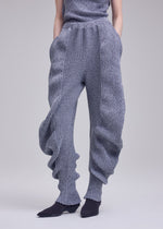 A person wears ISSEY MIYAKE's FOUND STONE chunky, textured knit pants with a mesh-like pattern, ribbed cuffs, and a voluminous silhouette, paired with black pointed shoes; upper body is partly out of frame against a plain background.