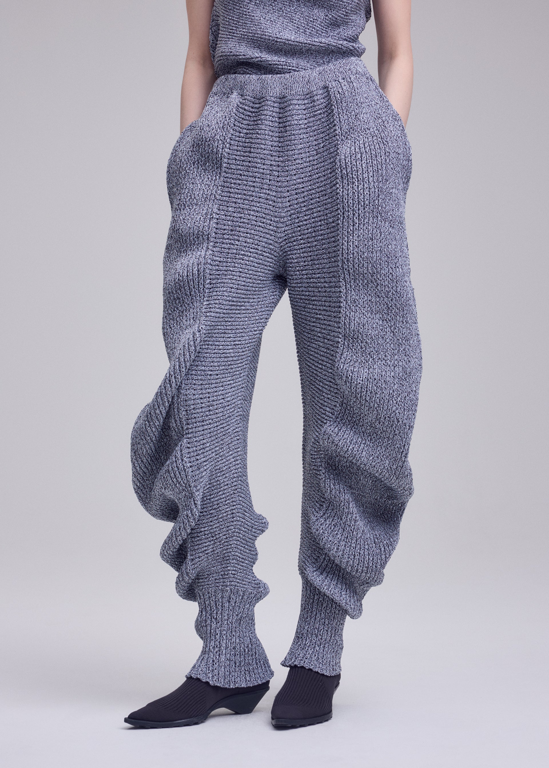 Zoomed view A person wears ISSEY MIYAKE's FOUND STONE chunky, textured knit pants with a mesh-like pattern, ribbed cuffs, and a voluminous silhouette, paired with black pointed shoes; upper body is partly out of frame against a plain background.