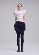 A woman models FOUND STONE by ISSEY MIYAKE, featuring a sculptural white sleeveless top with textured folds and a black sarouel skirt with exaggerated pockets, paired with black leggings and sock-like shoes. She faces the camera.
