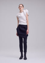A woman models the ISSEY MIYAKE FOUND STONE look: a white sculptural top with rounded shoulders and high neckline, paired with black sarouel pants featuring exaggerated pockets, plus matching black socks or shoes.