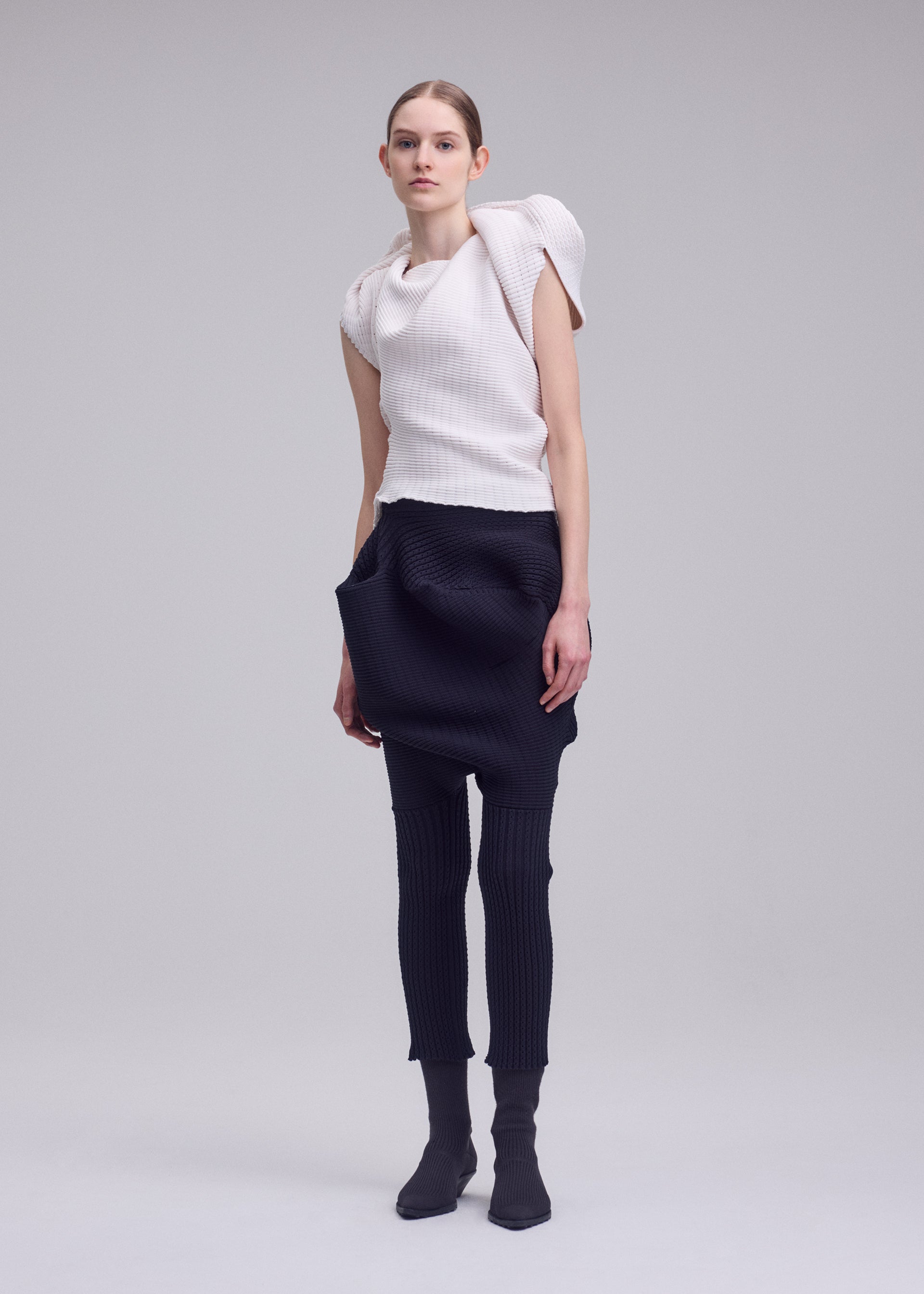 Zoomed view A woman models the ISSEY MIYAKE FOUND STONE look: a white sculptural top with rounded shoulders and high neckline, paired with black sarouel pants featuring exaggerated pockets, plus matching black socks or shoes.