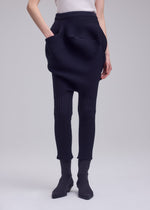 A person wears the FOUND STONE textured black skirt with large pockets by ISSEY MIYAKE over mesh pants and black sock boots, posing against a plain light background. The sculptural outfit stands out for its unique design.