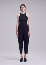 A person stands facing forward against a plain, light gray background wearing ISSEY MIYAKE’s CAMPAGNE ensemble: a sleeveless black top crafted from stretchy fabric, paired with high-waisted cropped black pants, and finished with black slip-on shoes.