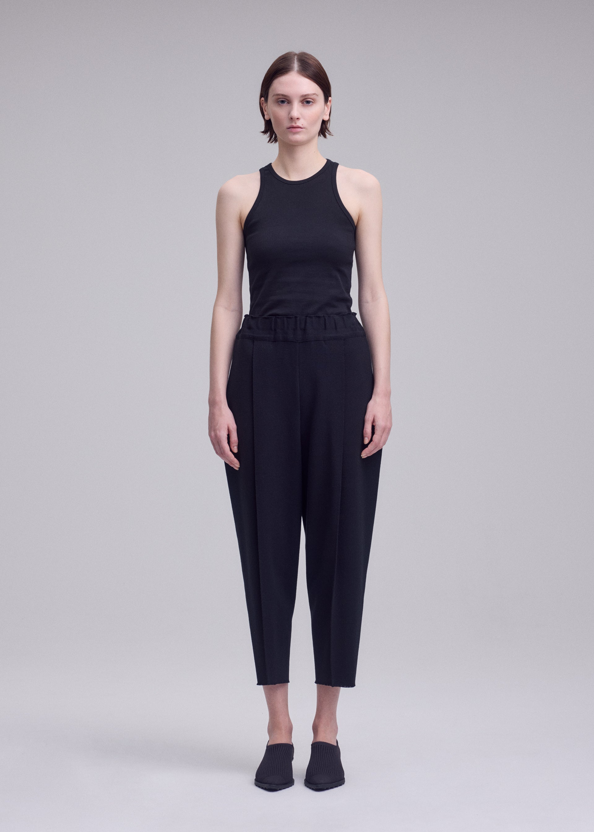 Zoomed view A person stands facing forward against a plain, light gray background wearing ISSEY MIYAKE’s CAMPAGNE ensemble: a sleeveless black top crafted from stretchy fabric, paired with high-waisted cropped black pants, and finished with black slip-on shoes.