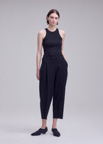 A person stands against a plain background, wearing ISSEY MIYAKE’s CAMPAGNE sleeveless black top and high-waisted, loose-fit cropped pants crafted from stretchy fabric, paired with black slip-on shoes. Their hands are in their pockets.