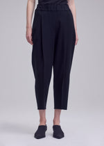 A person stands against a plain, light background wearing the ISSEY MIYAKE CAMPAGNE black high-waisted, cropped pants, crafted from stretchy fabric with a ruffled waistband, paired with black slip-on shoes. The upper body is partially out of frame.