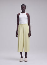 A person stands facing forward against a plain light gray background, wearing ISSEY MIYAKE’s CAMPAGNE light yellow wide pants crafted from soft, stretchy A-POC material, paired with a white sleeveless top and beige slip-on shoes.