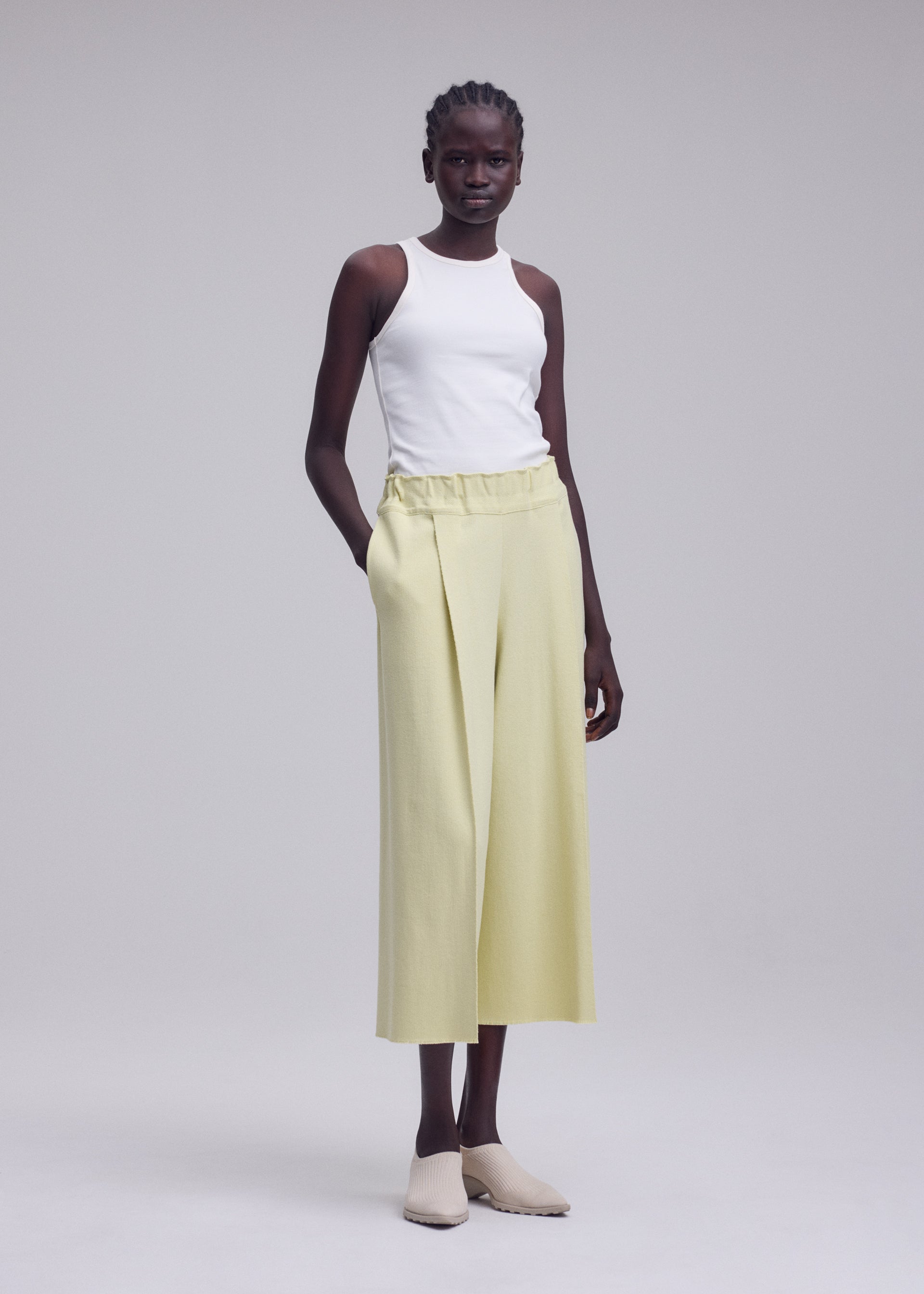 Zoomed view A woman stands with her hands in her pockets, wearing ISSEY MIYAKE’s CAMPAGNE light yellow wide-leg cropped pants crafted from innovative A-POC material, styled with a white sleeveless top and beige slip-on shoes against a plain light grey background.