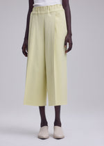 A person stands wearing ISSEY MIYAKE’s CAMPAGNE wide-leg cropped pants in light yellow A-POC material, paired with a white top and beige slip-on shoes against a plain light background.