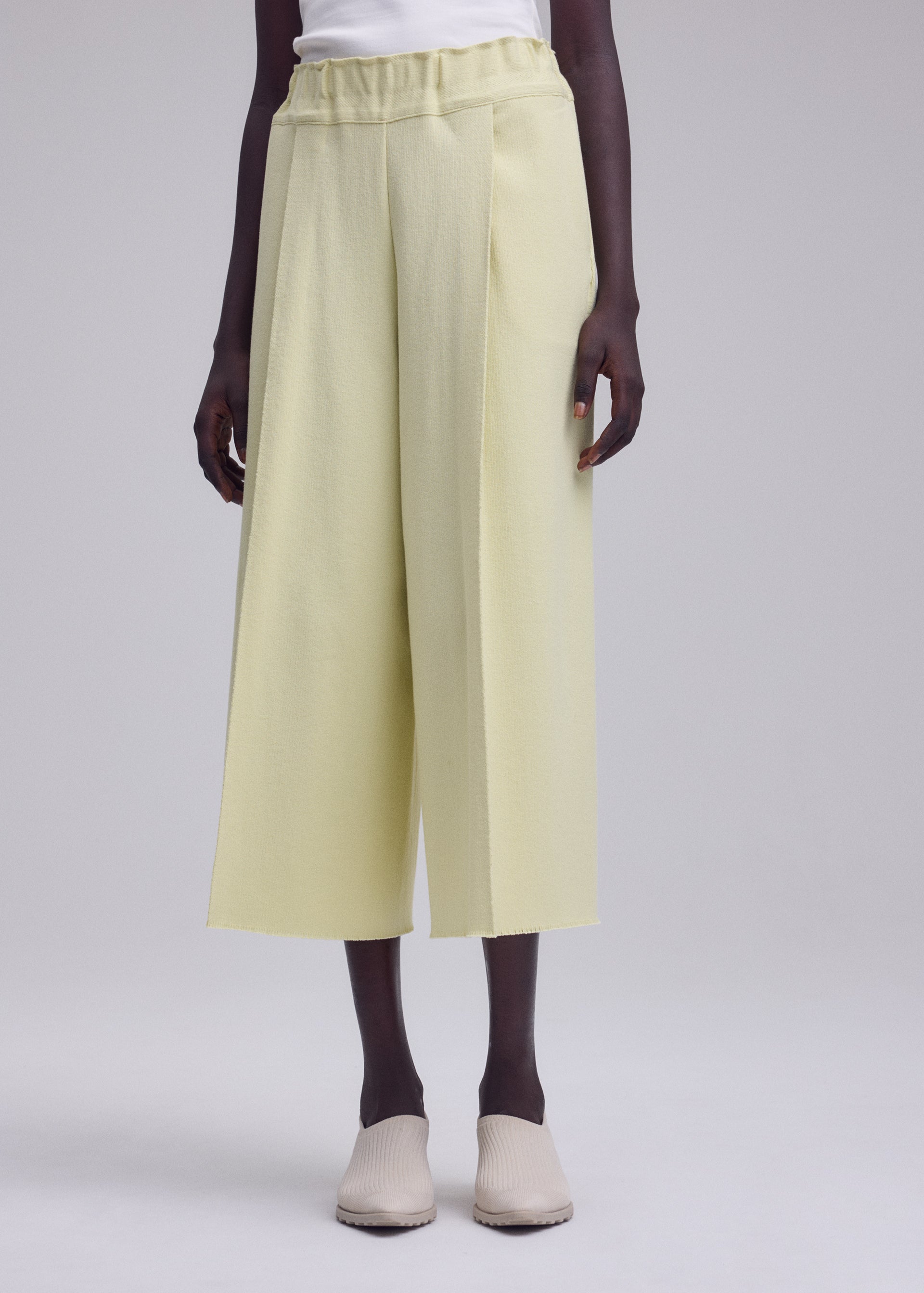 Zoomed view A person stands wearing ISSEY MIYAKE’s CAMPAGNE wide-leg cropped pants in light yellow A-POC material, paired with a white top and beige slip-on shoes against a plain light background.