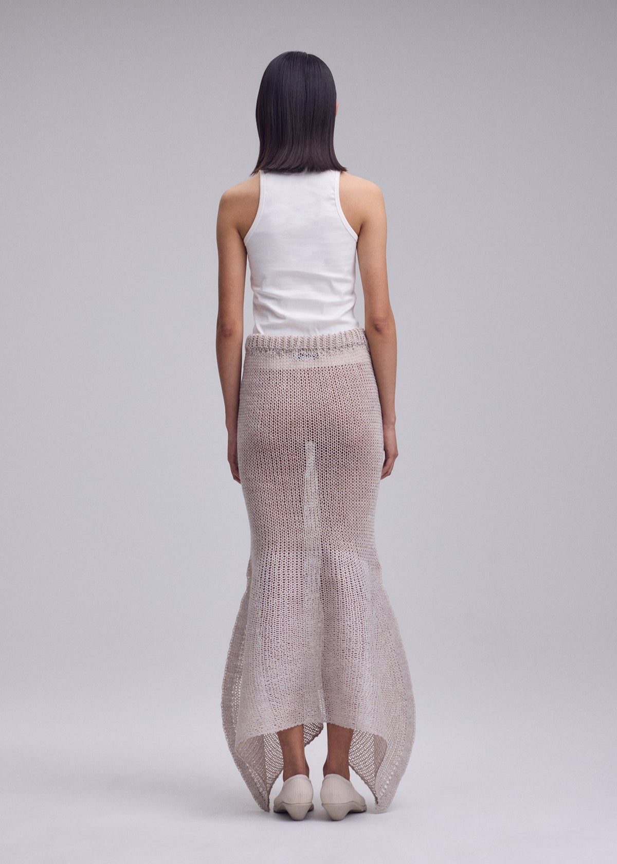 A person with straight, shoulder-length dark hair wears the ISSEY MIYAKE PANINI PULLED wool-alpaca knit skirt with a flared hem, a white sleeveless top, and white shoes against a plain light gray background. view 7 of 8