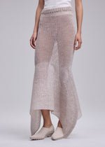 Person wearing a white sleeveless top, light beige shoes, and the PANINI PULLED skirt by ISSEY MIYAKE—sheer wool-alpaca knit with an uneven hem—stands against a plain light gray background.