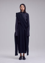 A woman faces forward in a dark, asymmetrical ISSEY MIYAKE outfit featuring the O KNIT 100% wool skirt with long sleeves and a draped scarf. She has straight dark hair and a neutral expression against a light gray background.