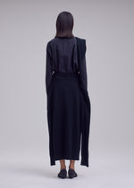 Person stands with their back to the camera, wearing a long-sleeved black shirt, ISSEY MIYAKE O KNIT skirt, and black shoes against a plain light gray background.