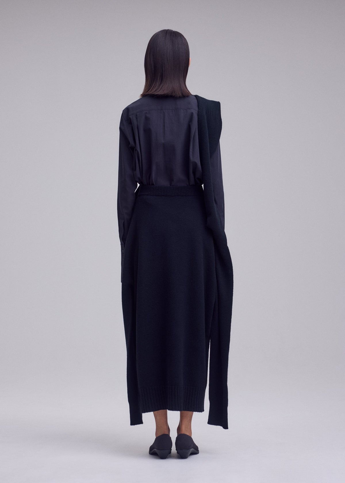 Zoomed view Person stands with their back to the camera, wearing a long-sleeved black shirt, ISSEY MIYAKE O KNIT skirt, and black shoes against a plain light gray background.