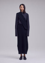 A person stands facing forward in a long, dark, minimalist ISSEY MIYAKE outfit featuring the unique asymmetrical 100% wool O KNIT overlay, with dark shoes and straight hair, against a plain light gray background.