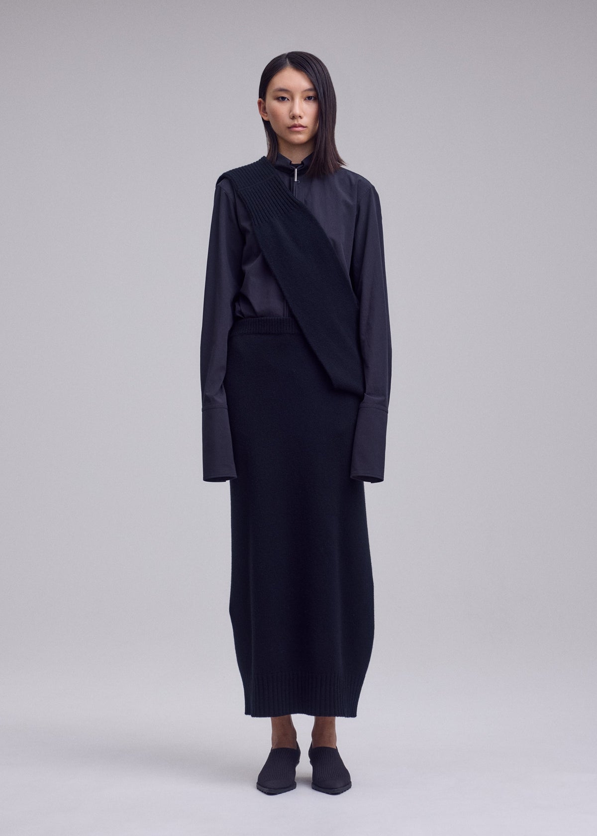 Zoomed view A person stands facing forward in a long, dark, minimalist ISSEY MIYAKE outfit featuring the unique asymmetrical 100% wool O KNIT overlay, with dark shoes and straight hair, against a plain light gray background.