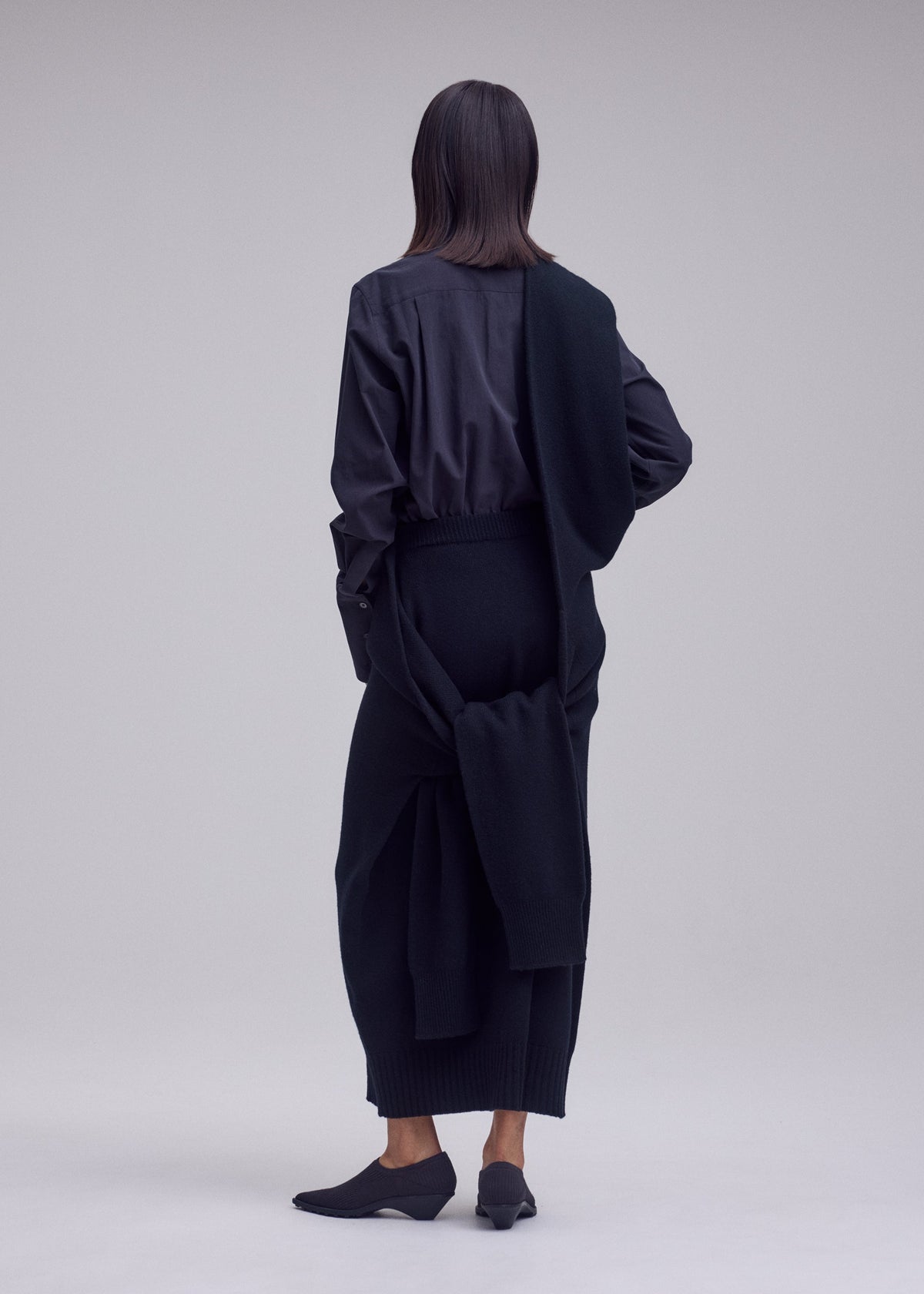 Zoomed view A person with straight shoulder-length hair faces away, wearing ISSEY MIYAKE’s O KNIT as a long skirt, a dark blouse, matching shoes, and a sweater draped over their shoulder and tied at the waist against a plain light background.