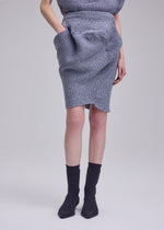 Wearing the ISSEY MIYAKE FOUND STONE knit mini skirt with side pockets and a matching top, paired with black ankle socks and sock-style shoes, a person stands against a plain light gray background.