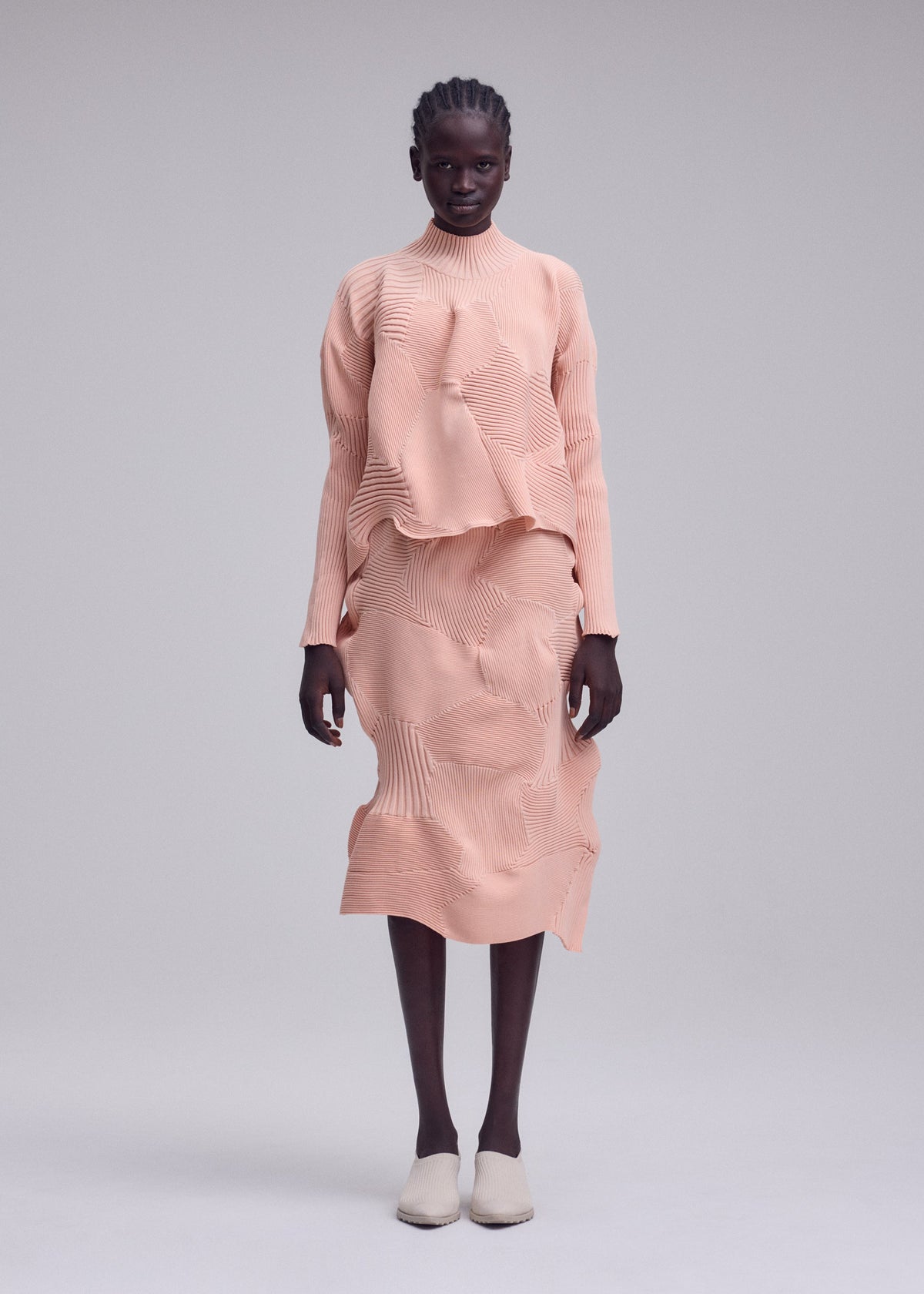 A person stands against a light gray background, wearing the KONE KONE textured peach-colored long-sleeve top and matching knit skirt with uneven texture and wave-like patterns by ISSEY MIYAKE, complemented by beige slip-on shoes. view 4 of 6