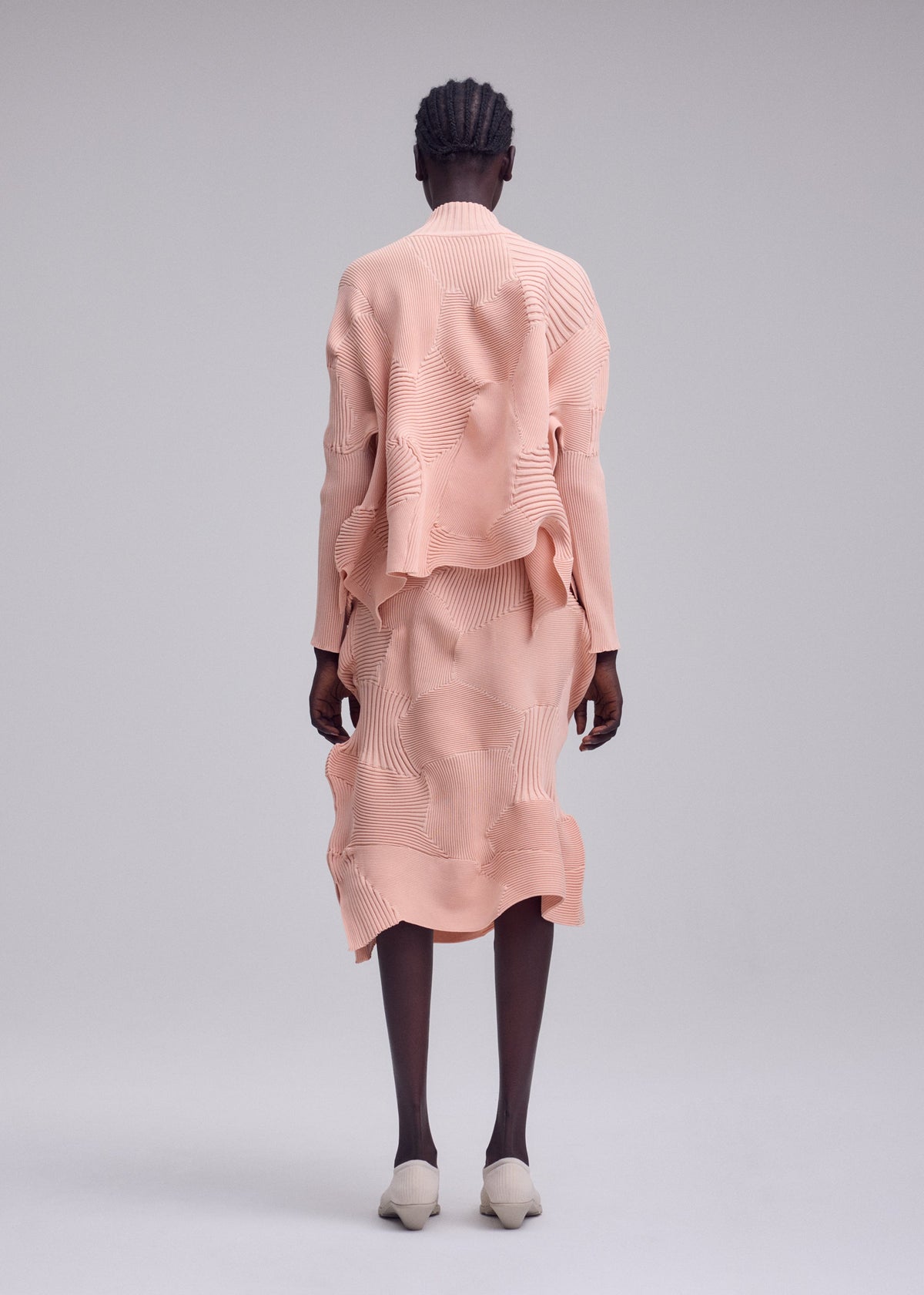 Someone stands with their back to the camera, wearing the ISSEY MIYAKE KONE KONE set—a textured, peach-toned outfit consisting of a knit skirt and long-sleeved top. Their hair is braided, and they wear light shoes while posing against a plain gray backdrop. view 5 of 6