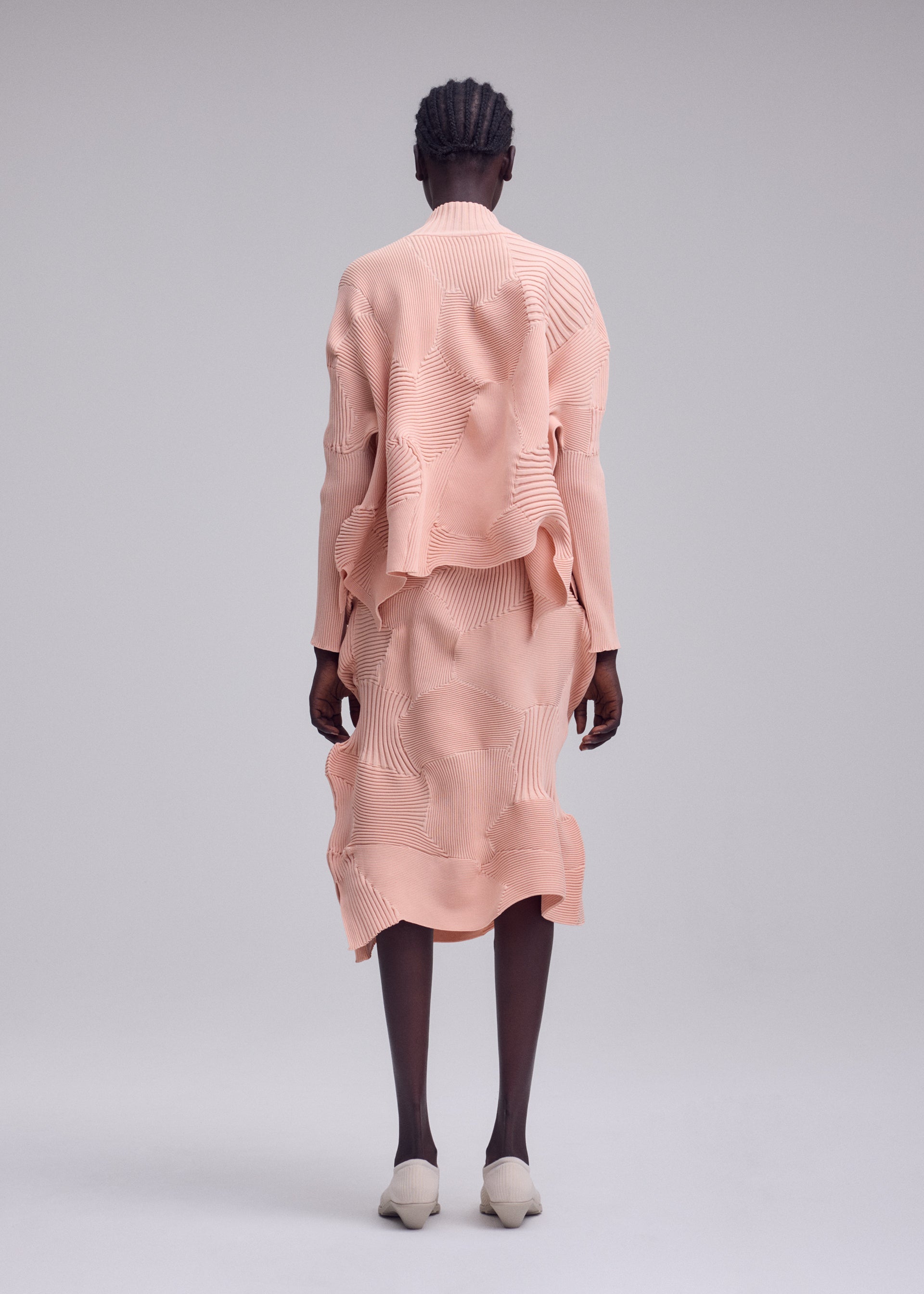 Zoomed view Someone stands with their back to the camera, wearing the ISSEY MIYAKE KONE KONE set—a textured, peach-toned outfit consisting of a knit skirt and long-sleeved top. Their hair is braided, and they wear light shoes while posing against a plain gray backdrop.