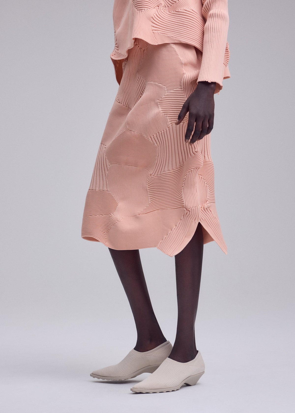 A person wearing the ISSEY MIYAKE KONE KONE textured light peach knit skirt with a subtle linear pattern and a matching top stands against a plain background. They pair the outfit with light beige slip-on shoes, with only the lower half of their body visible. view 6 of 6