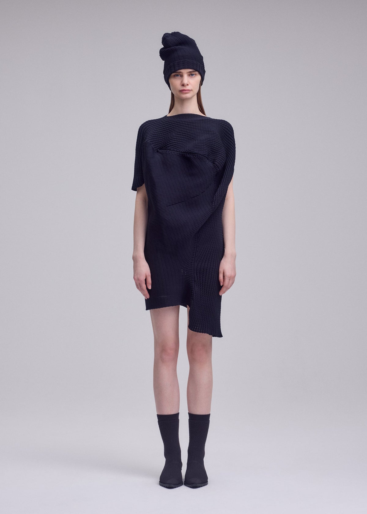 A woman stands against a light gray background, wearing the FOUND STONE textured black mini dress by ISSEY MIYAKE with an asymmetrical shape, paired with a matching black beanie, black ankle socks, and pointed shoes. She looks straight ahead, expression neutral. view 5 of 6
