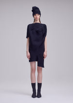 A woman stands against a light gray background, wearing the FOUND STONE textured black mini dress by ISSEY MIYAKE with an asymmetrical shape, paired with a matching black beanie, black ankle socks, and pointed shoes. She looks straight ahead, expression neutral.