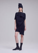 A woman stands against a plain background wearing the FOUND STONE asymmetrical black knit dress and matching tall hat by ISSEY MIYAKE, paired with black socks and pointed-toe shoes. She looks ahead with a neutral expression.