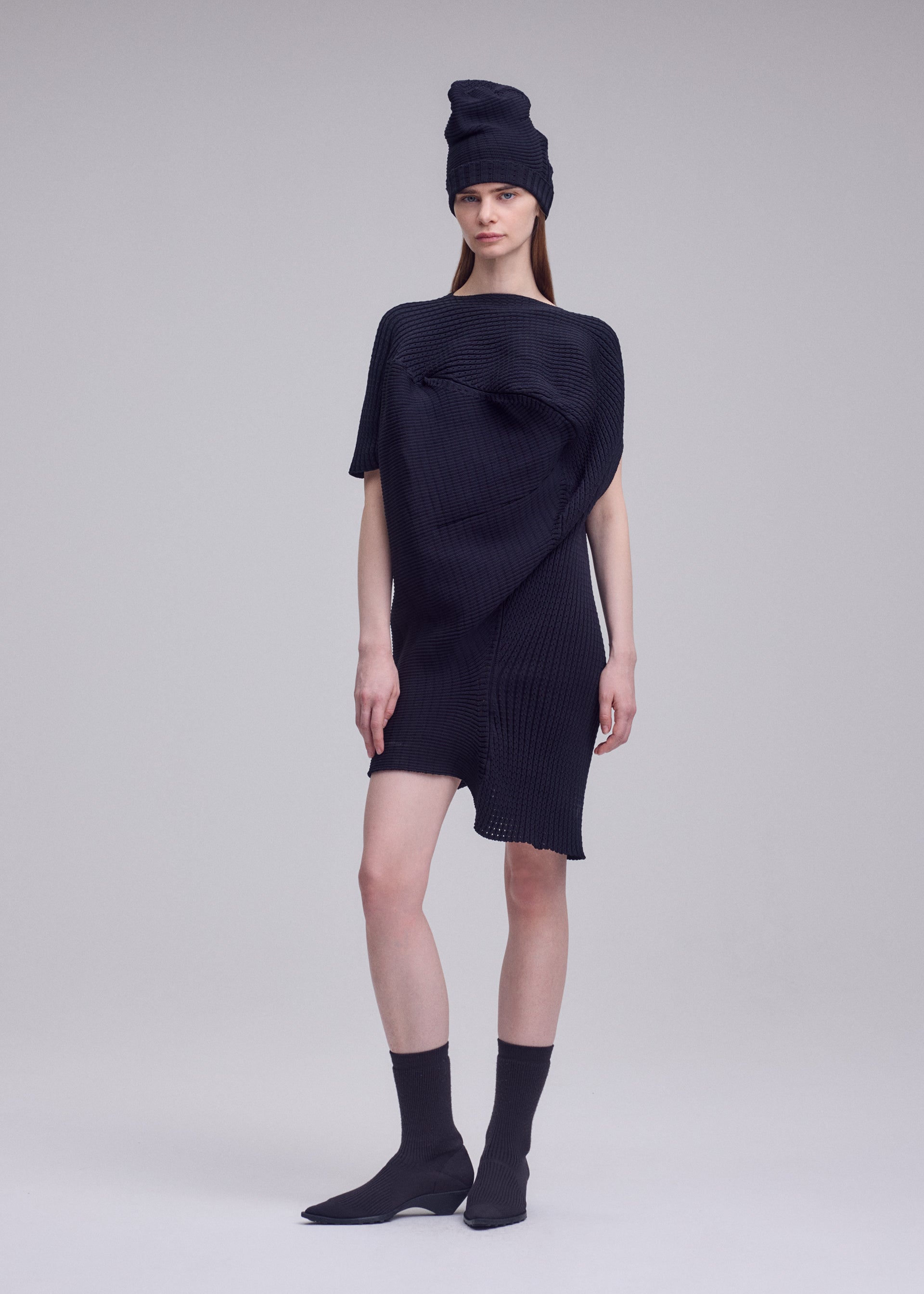 Zoomed view A woman stands against a plain background wearing the FOUND STONE asymmetrical black knit dress and matching tall hat by ISSEY MIYAKE, paired with black socks and pointed-toe shoes. She looks ahead with a neutral expression.