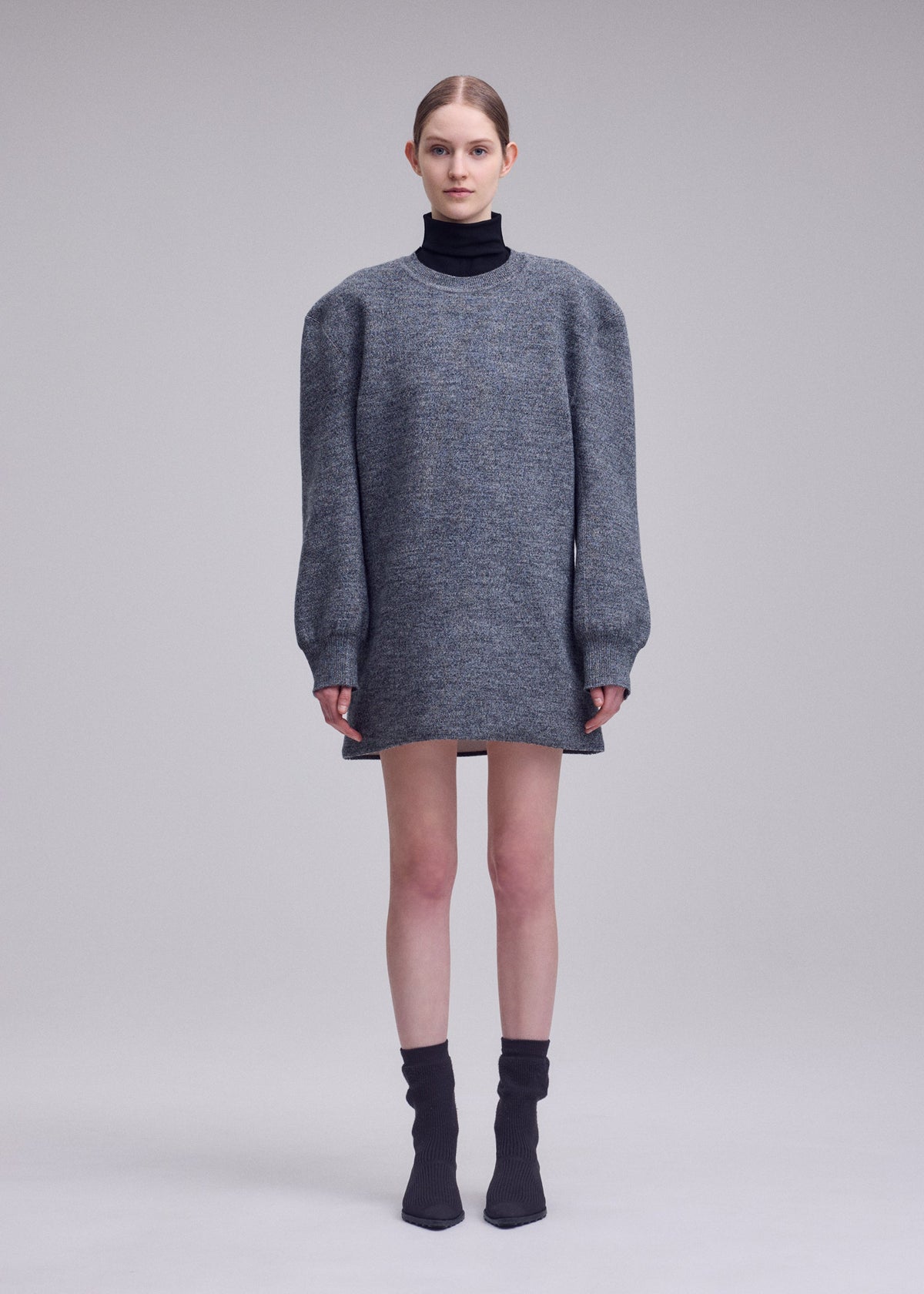 Zoomed view A woman wears the ISSEY MIYAKE HANDSOME KNIT gray mini dress with structured shoulders and long sleeves, styled with a black turtleneck, black socks, and pointed shoes against a plain light background.