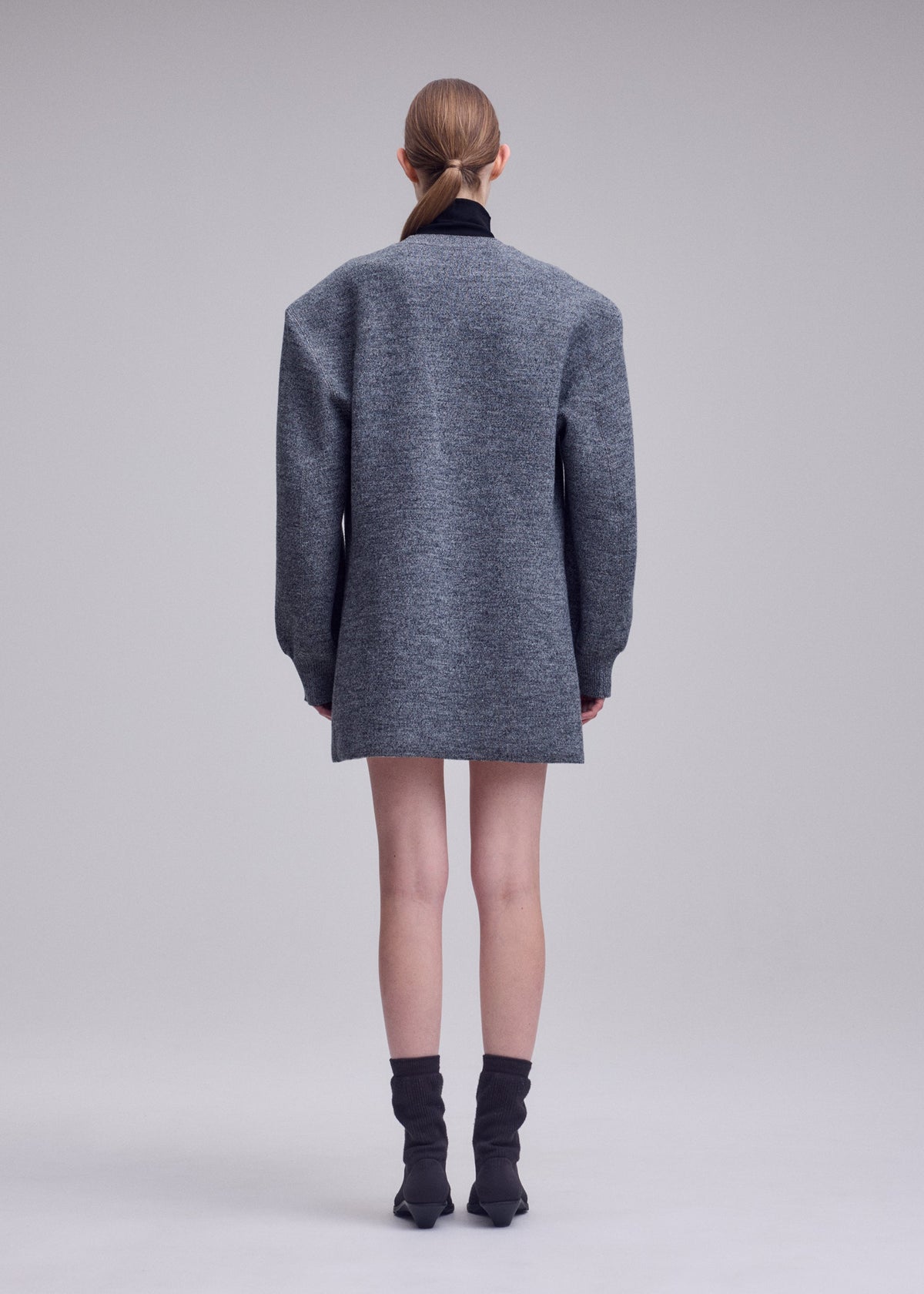 A person stands facing away in a long gray HANDSOME KNIT by ISSEY MIYAKE, black socks, and pointed shoes. Their hair is tied back tightly against a plain light background. view 5 of 6