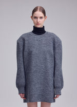 A person with light skin and brown hair tied back wears a long, oversized gray HANDSOME KNIT by ISSEY MIYAKE over a black turtleneck, standing against a plain light gray background.