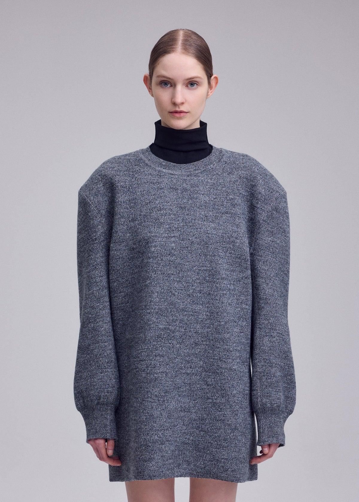 Zoomed view A person with light skin and brown hair tied back wears a long, oversized gray HANDSOME KNIT by ISSEY MIYAKE over a black turtleneck, standing against a plain light gray background.